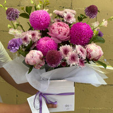 A vibrant flower box featuring bright pink and magenta chrysanthemums, light pink peonies, purple pompons, and white chrysanthemums, wrapped in white tissue paper and a white box with a purple ribbon