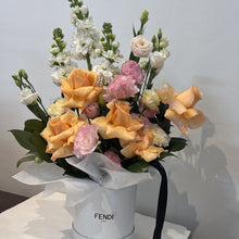 A floral arrangement in a white cylindrical box, featuring peach roses, pink flowers, white blooms, and green foliage, 