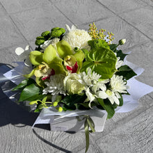 A fresh flower bouquet featuring green orchids, white chrysanthemums, and green foliage, wrapped in white paper and placed on a gray paved surface.